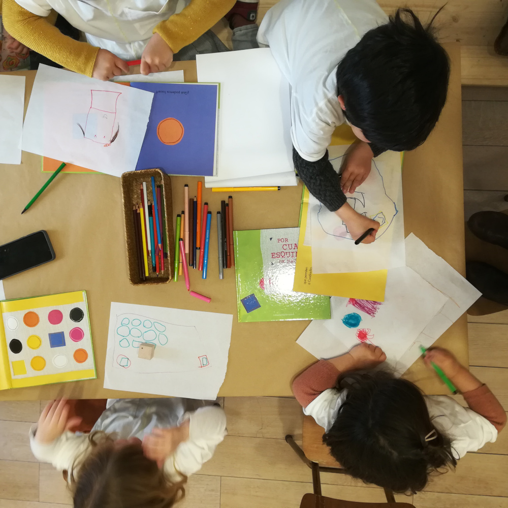 Vista superior de un grupo de niñas y niños pintando sobre una mesa con lápices, folios y libros.